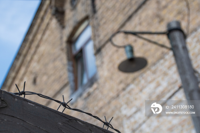 Close-up of a rusty barbed wire fence surrounding a concentration and extermination camp, old barrack with window and lamp in soft focus in the background