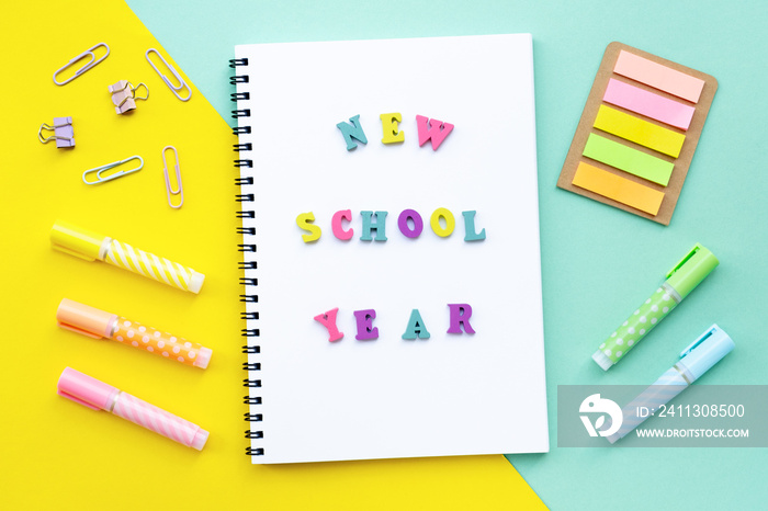 New school year inscription made from multicolored letters. Stack of spiral notebooks overlap. Colorful yellow with mint color background. Markers and markers and other office supplies around