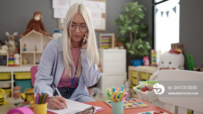 Young blonde woman preschool teacher talking on smartphone writing on document at kindergarten