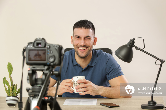 Young smiling cheerful man sitting at the office desk in front of the camera making a video. Male blogger recording a video tutorial.