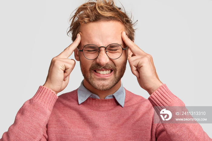 Miserable male clenches teeth from tiredness and pressure on work, keeps index fingers on temples, wears sweater, tries to gather with thoughts, isolated over white studio wall. Facial expressions