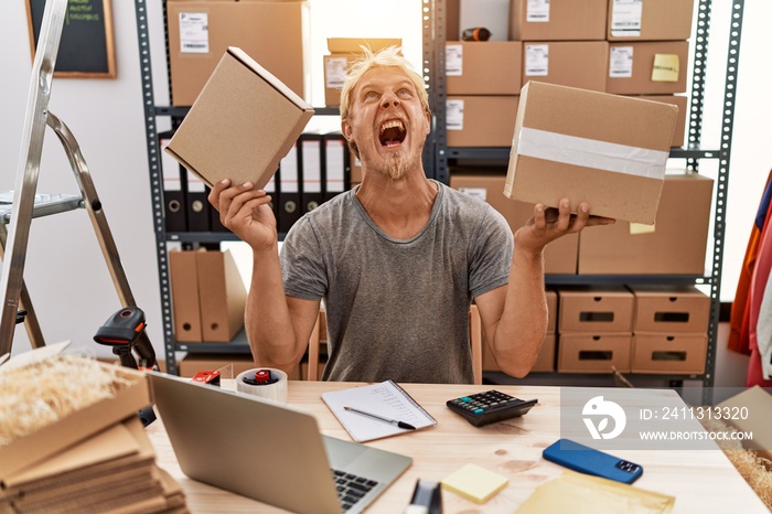 Young blond man holding packages working at online shop angry and mad screaming frustrated and furious, shouting with anger looking up.