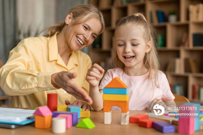 Cute little girl and cheerful child development specialist playing with colorful wooden bricks, sitting at table