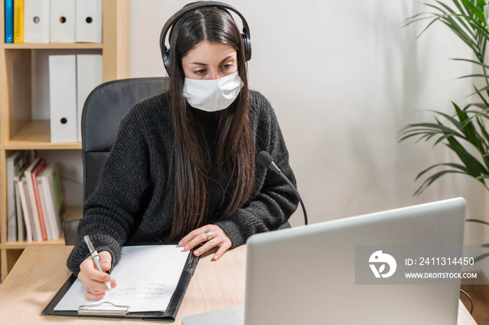 Telemarketer woman working on laptop and talking on a headset with face mask at office. Self employed and working from home because of coronavirus. Business worker answer customer calls.