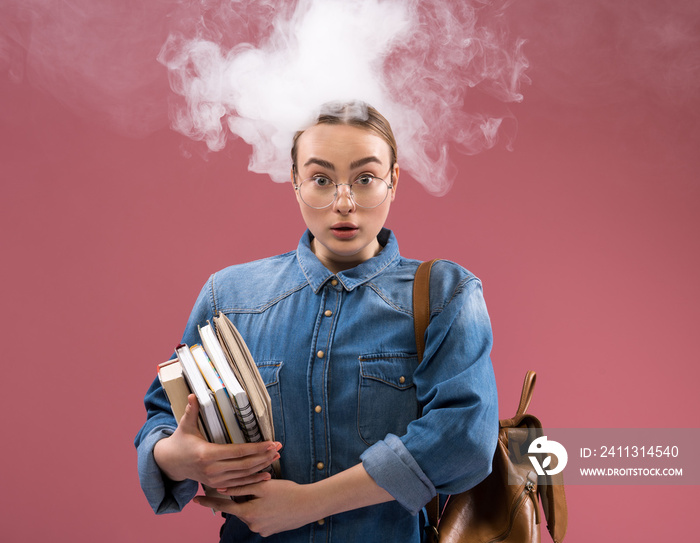 Too much homework. Portrait of smart young woman having her brain smoked. She is looking at camera with shock while keeping volumes in hands