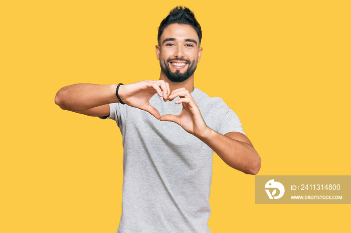 Young man with beard wearing casual grey tshirt smiling in love doing heart symbol shape with hands. romantic concept.