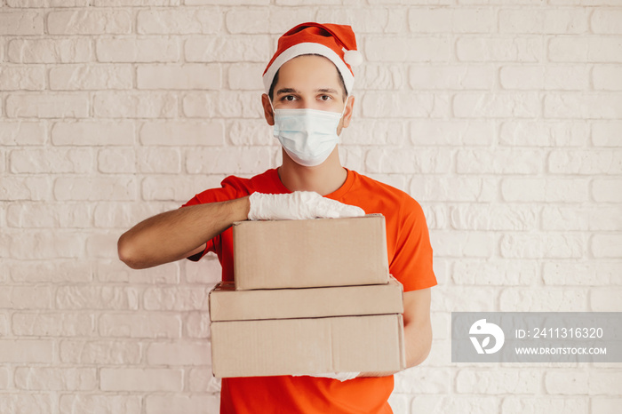 Young man courier in protective face mask, medical gloves, santa hat carry delivery boxes in hands. Happy delivery service employee in medical face mask hold cardboard packages. Christmas, New Year