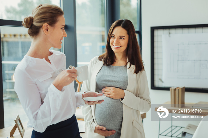 Drinking coffee. Pretty cute pregnant woman standing with her hand on a belly and smiling while looking at her cheerful emotional coworker