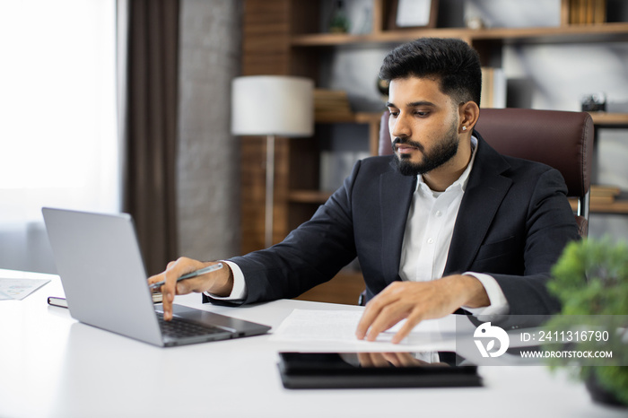 Confident businessman wearing suit writing notes or financial report, sitting at desk with laptop, focused serious man working with paper documents, student studying online, research work