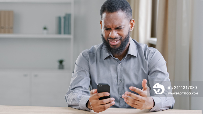 Angry African American ethnic bearded man boss businessman with mobile phone smartphone mistake failure reading bad news refuse notice mad with business trouble problem sitting at table in office