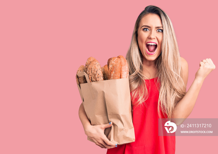 Young beautiful blonde woman holding paper bag with bread screaming proud, celebrating victory and success very excited with raised arms