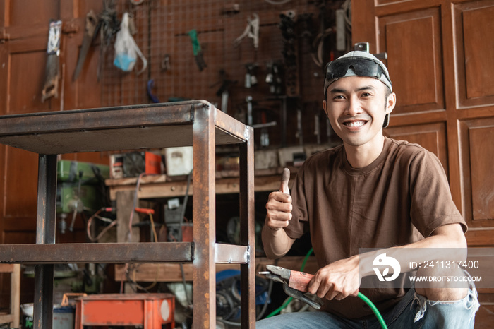 close up of Male welder smiling with a thumbs up while holding an electric welder in a welding workshop background