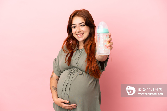 Young redhead caucasian woman isolated on pink background pregnant and holding a feeding bottle