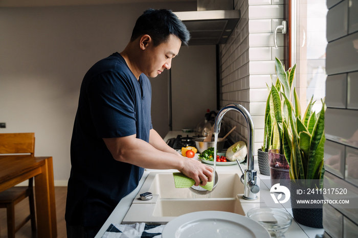 Adult brunette asian man wearing t-shirt washing dishes at kitchen
