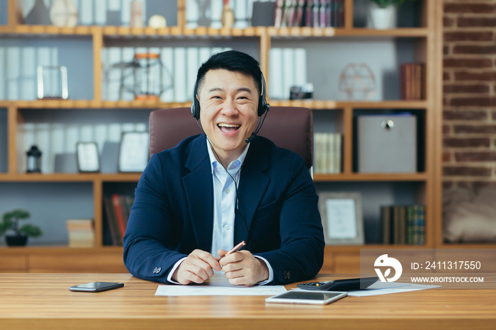 Happy Asian businessman boss looks at the camera smiling and talking to a colleague in an online conference, uses a headset, a man in a business suit works in the office