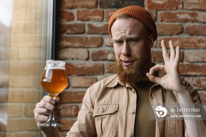 Bearded man drinking beer in pub. Positive hipster guy holding glass with alcohol, tasting beer quality and having fun, show ok sign