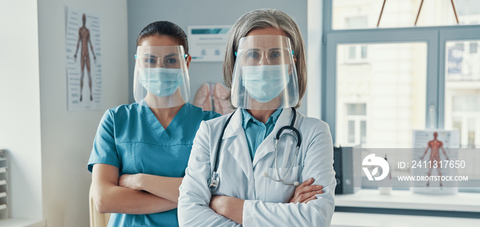 Two confident women coworkers in medical uniform and protective workwear keeping arms crossed and looking at camera while working in the hospital