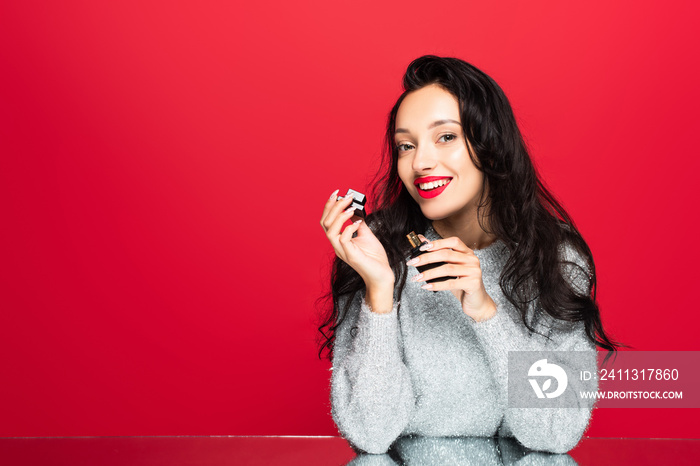 young pleased woman in sweater holding bottle with perfume isolated on red