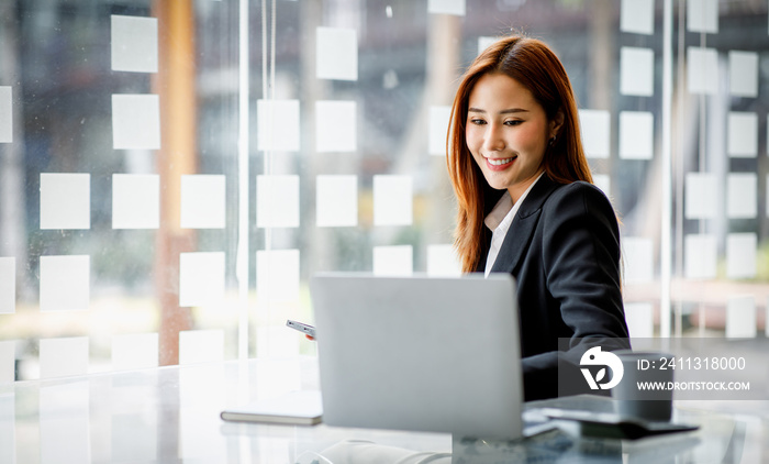 Photo of cheerful pretty Asian woman having been employed to a job as executive smiling toothily sitting at the desktop with laptop and writing note.