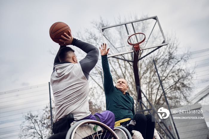 Basketball player in wheelchair taking a shot while playing with friend on outdoor court.