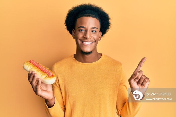 African american man with afro hair eating hotdog smiling happy pointing with hand and finger to the side