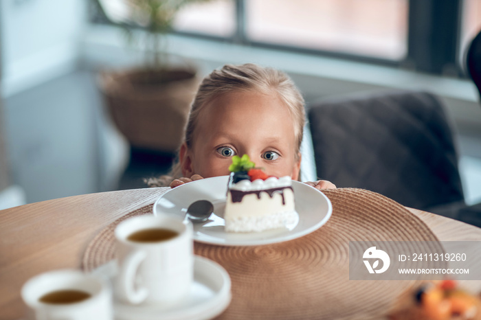 A cute blonde girl looking anticipated while looking at the slice of cake