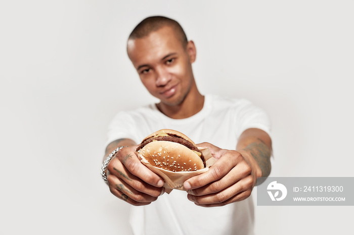 Tasty looking hamburger in hands of a dark-skinned tattooed man wearing white t-shirt, looking at camera and smiling on a white background