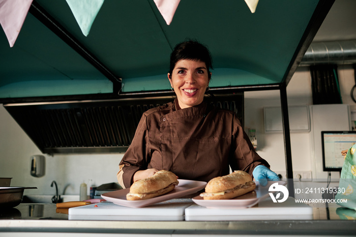 Woman looking at camera and smiling while serving two sandwiches at restaurant counter.