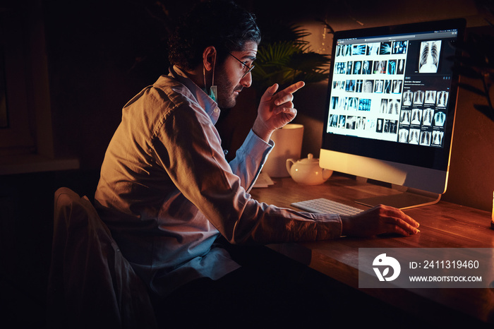 Smart medic in glasses is working with x-ray screens using a computer at dark office.