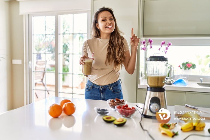 Beautiful young brunette woman drinking glass of smoothie at the kitchen surprised with an idea or question pointing finger with happy face, number one