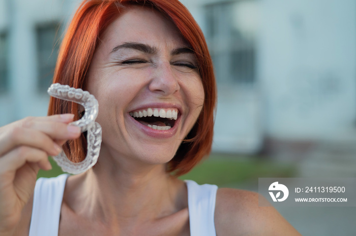 Red-haired Caucasian woman holding transparent mouthguards for bite correction outdoors. A girl with a beautiful snow-white smile uses silicone braces