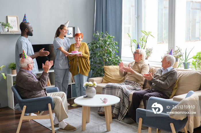 Nurse and doctor congratulating senior woman with her birthday and giving a cake to her with other senior people clapping hands