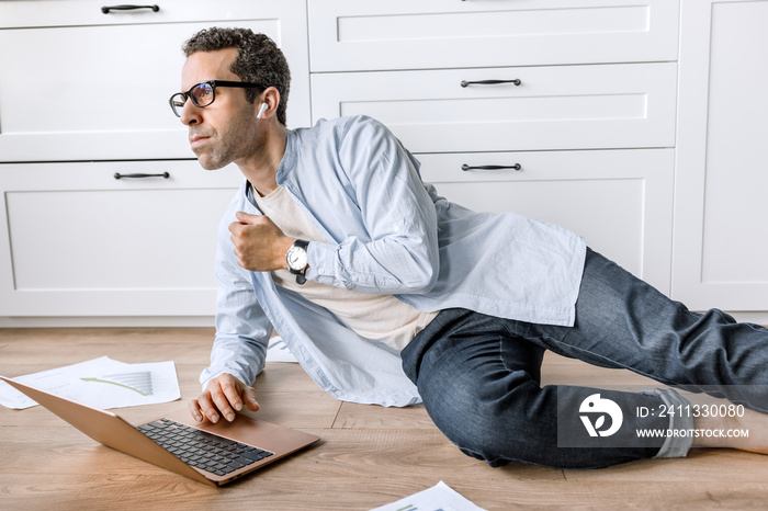 Concentrated businessman lays on floor at home, using laptop, developing new project. Thoughtful stylish male employee or student is working or studying remotely, learning financial graphs