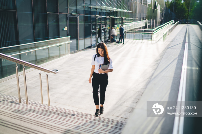 Happy Caucasian schooler with modern digital tablet in hand walking near college building located at city urbanity, carefree hipster girl in casual wear holding touch pad and smiling outdoors