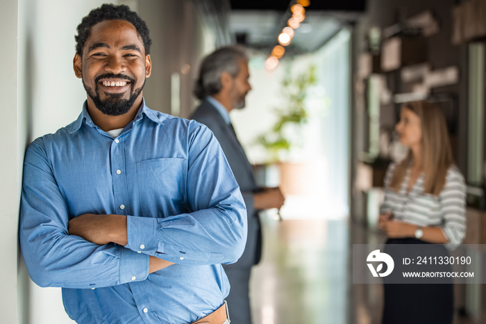 Portrait Of Businessman In Modern Open Plan Office With Business Team Working In Background. African office worker with arms crossed. Successful businessman in modern office