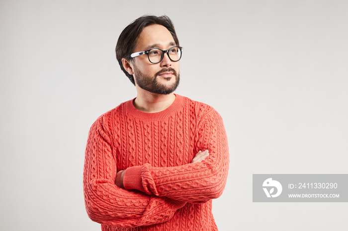 Content dreamy young Asian man with cool beard and mustache wearing warm knitted sweater crossing arms on chest