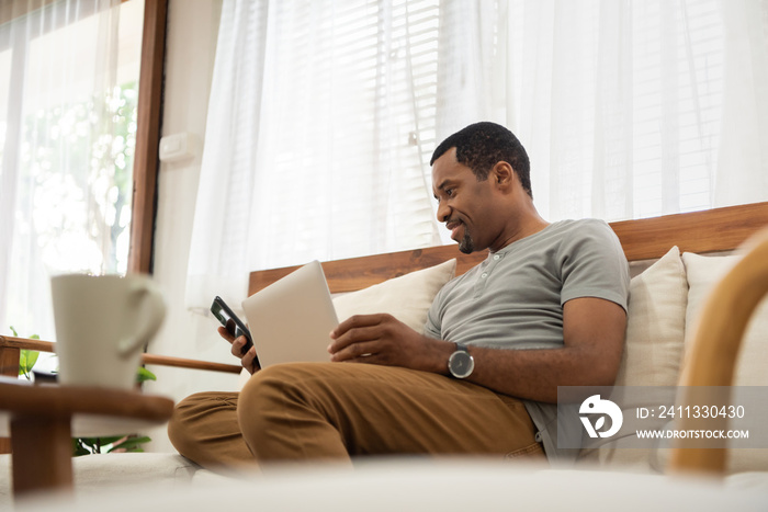 Smiling African American man using smartphone while sitting on sofa at home. Black Male Freelancer texting on mobile phone in Living room. Lifestyle concept.