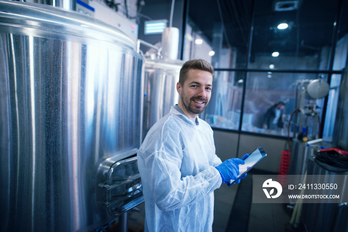 Portrait of caucasian man in white suit standing by the reservoirs in production plant. Industrial worker with tablet.