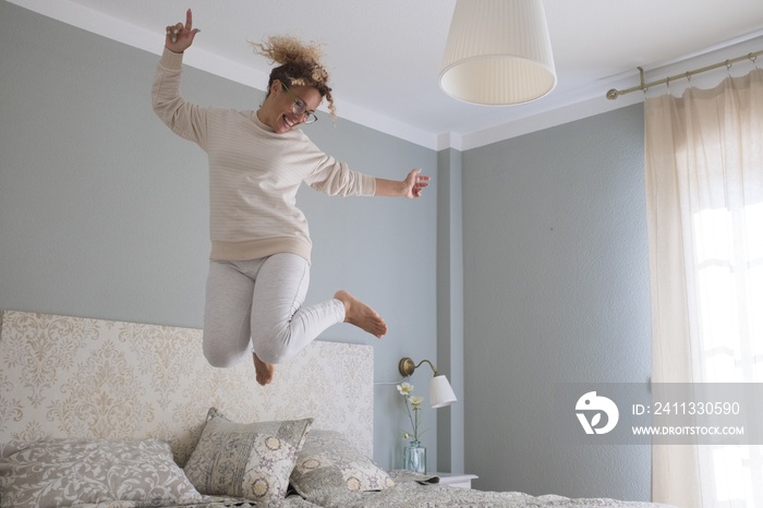 Crazy young woman jumping on bed at home. Overjoyed woman going crazy. Carefree woman dancing and jumping on bed in excitement celebrating success at modern apartment bedroom