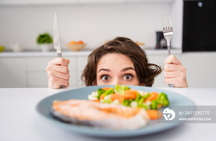 Close up photo of housewife lady cunning tricky hungry eyes look from under table ready to eat grilled salmon trout fillet steak garnish portion hold fork knife modern kitchen indoors