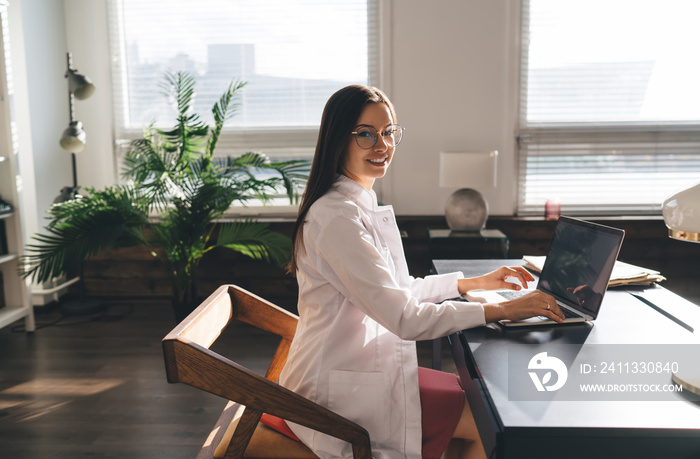 Cheerful female physician in medical uniform typing on computer