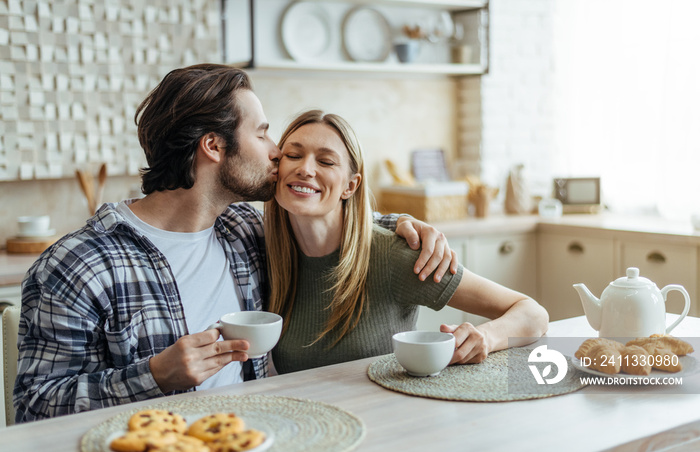 Smiling millennial caucasian husband with stubble hugs and kisses his wife on cheek, couple drinks coffee