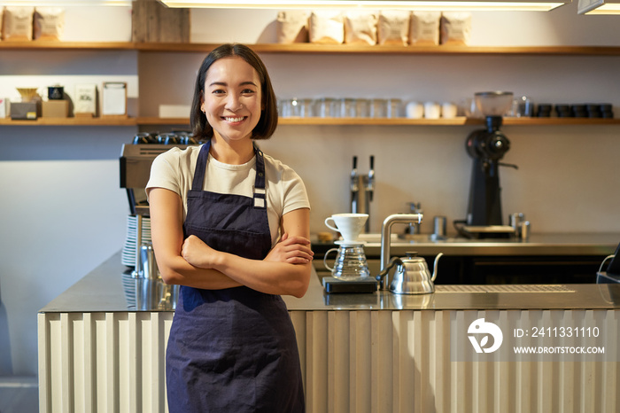 Portrait of smiling asian female barista, wearing apron, standing near counter with coffee, working in cafe