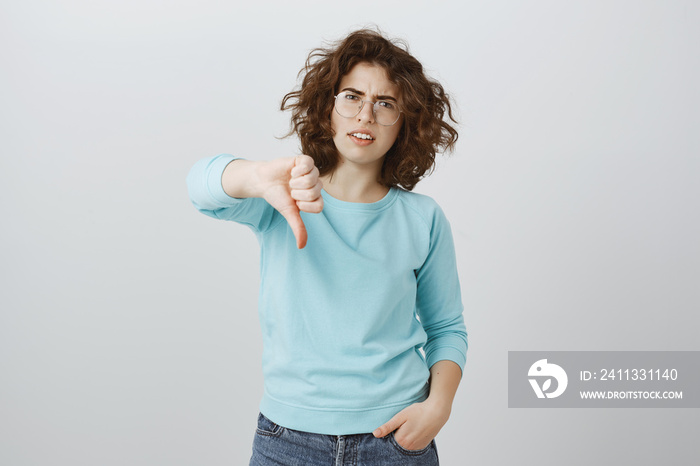 Girl displeased with bad idea. Indoor shot of annoyed displeased young woman with curly hair in trendy glasses, showing thumbs down, frowning and expressing dislike, rejecting plan over gray wall