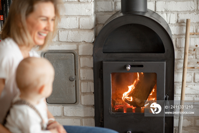 Portrait of young smiling blond woman with baby in hands basking near comfort warm fireplace with firewood. Side view