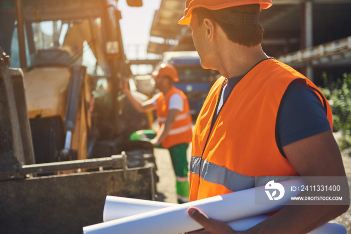 Waist up of adult worker is holding drafts in hands while his colleague standing near excavator on backgroung