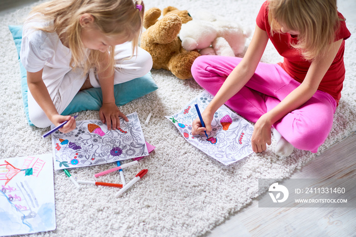 High angle portrait of two little sisters coloring pictures together sitting on thick carpet at home in cozy living room