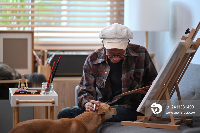 Smiling asian man painting picture in living room and playing with his dog.