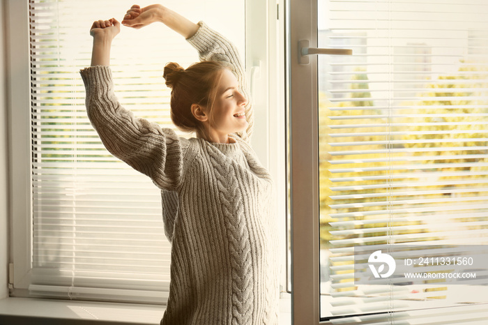 Happy young girl stretching near window at home