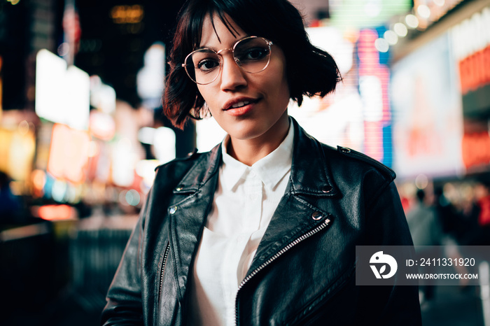 Portrait of gorgeous brunette hipster girl with stylish short haircut looking at camera on night city background with neon illumination.Charming female in eyewear and trendy jacket standing on street